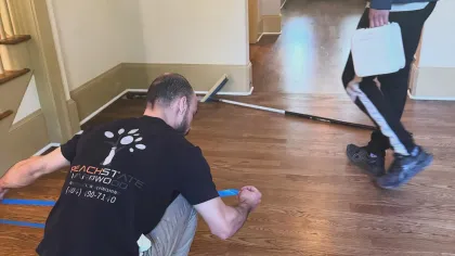 Worker applying blue painter's tape on hardwood floor in residential hallway for finishing or protection.
