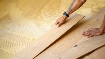 Hands installing wooden floor planks over adhesive with tattooed arm and smartwatch visible