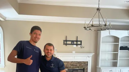 Two workers installing hardwood flooring in a bright room with fireplace, shelves, and chandelier