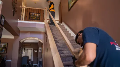 Two workers installing a wooden handrail on a residential carpeted staircase inside a home with brown walls.