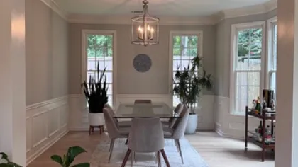 Modern dining room with glass table, gray chairs, plants, chandelier, light wood flooring, and large windows.