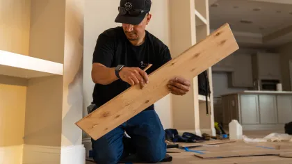 Man in black shirt and cap measuring wooden plank while installing flooring in home interior.