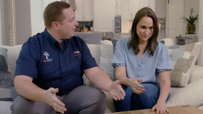 Man and woman sitting on a couch discussing while gesturing over a wooden table in a living room.