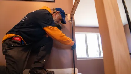 Worker wearing a cap and gloves installing or repairing wooden stair railing indoors near a window