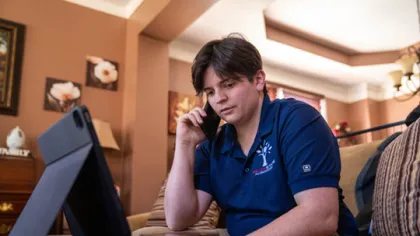 Teenage boy in blue polo shirt using a phone and tablet at home on the couch with brown walls and framed art.
