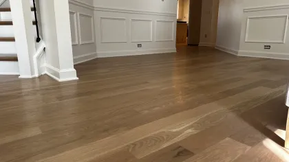 Spacious room with polished hardwood floors, white paneled walls, and staircase with wooden steps and black railing.