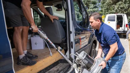 Two men unloading a floor buffer machine from a van on a sunny day outdoors.