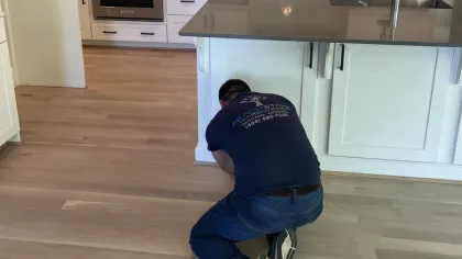Worker kneeling on light hardwood floor in modern white kitchen with island and stainless steel appliances.