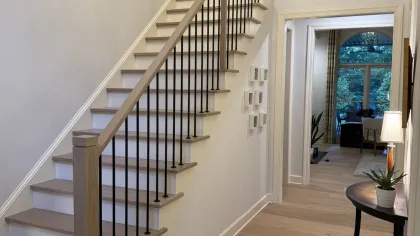 Modern wooden staircase with black metal balusters in a bright hallway leading to a living room with large window.