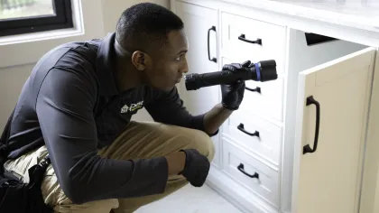 Home inspector examining cabinet inside a kitchen with flashlight, wearing gloves and uniform.