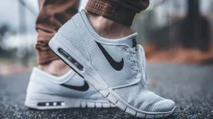 Close-up of white Nike sneakers with black swoosh worn with brown pants on asphalt ground outdoors.