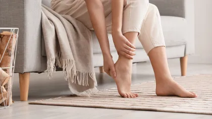 Person sitting on gray sofa holding their foot, indicating foot pain or discomfort in a cozy living room.