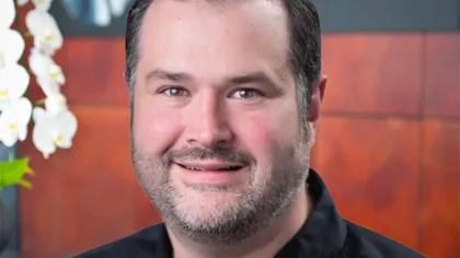 Smiling man with short dark hair and beard wearing black shirt in front of Pirtek logo and white flowers.