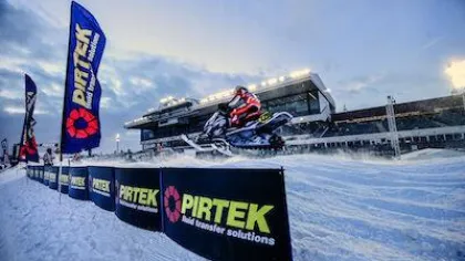 Snowmobile rider in red gear jumps over snowy track at a Pirtek sponsored winter race event with grandstand in background