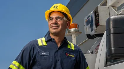 Smiling industrial worker in blue uniform and yellow hard hat with machinery in the background under clear sky