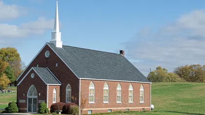 Red brick church with white steeple in a grassy rural area under a partly cloudy blue sky.