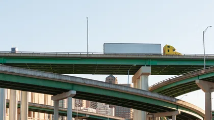 Yellow semi-truck driving on elevated green highway overpasses with city skyline in the background on clear day