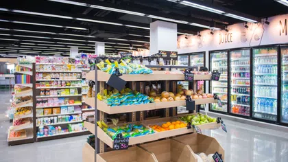 Bright supermarket aisle with fresh fruits, snacks on wooden shelves, and refrigerated drinks in the background.