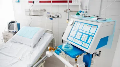 Hospital ICU room with empty bed and advanced mechanical ventilator with blue and white controls.
