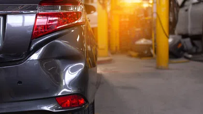 Close-up of a gray car with a dented rear bumper parked inside an auto repair shop with bright yellow pillars.