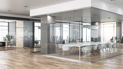 Modern office with glass meeting room, wooden flooring, white chairs, and natural light from large windows.