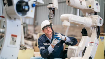 Technician wearing safety gear repairs a white industrial robotic arm inside a factory environment.