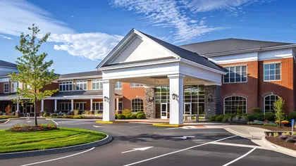 Modern brick building with white entrance canopy, landscaped greenery, and a circular driveway under blue sky.