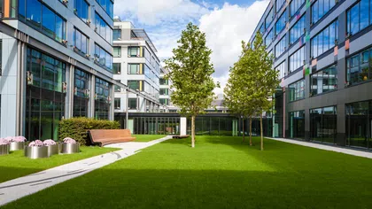 Modern office courtyard with green lawn, trees, benches, and glass buildings under a partly cloudy sky
