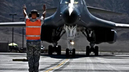Ground crew in orange vest guiding a large military aircraft on the runway with mountains in the background