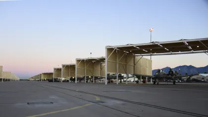 Row of military aircraft parked under protective canopies at an airbase with mountains in the background at dusk