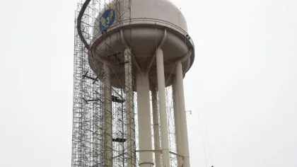 Tall water tower surrounded by scaffolding with workers performing maintenance against a gray sky.