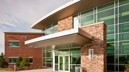 Modern office building entrance with glass windows, stone facade, and red brick side wall under clear sky.