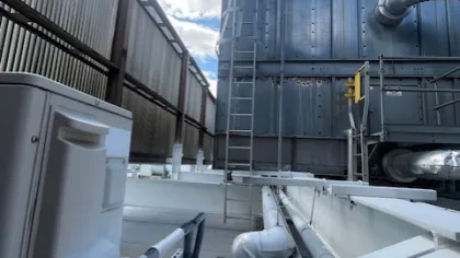 Industrial rooftop with metal pipes, ventilation ducts, and a ladder under a partly cloudy sky.