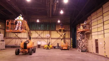 Workers using orange aerial lift platforms inside a large industrial warehouse with high ceilings.