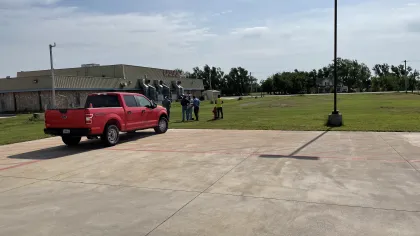 Red pickup truck parked near a building with four people standing on a grassy field under a clear sky.