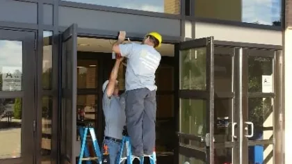 Two workers install or repair a glass entrance door at a commercial building using ladders and safety cones.