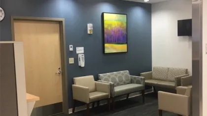 Modern waiting area with beige chairs, dark blue accent wall, colorful painting, and mounted TV in office space
