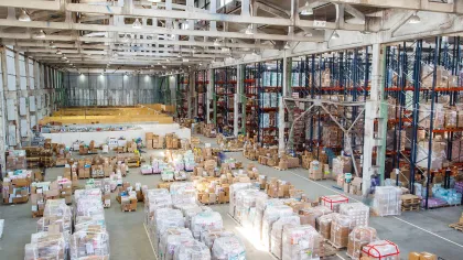 Large warehouse interior filled with pallets of boxed goods, organized on shelves and open floor space.