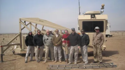 Group of men standing in front of beige military equipment in a desert setting under clear sky.