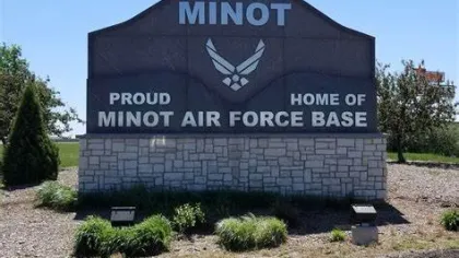 Stone sign reading Minot Proud Home of Minot Air Force Base with Air Force logo and greenery under blue sky