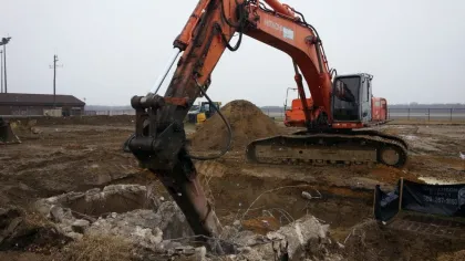 Orange excavator with hydraulic breaker demolishing concrete on a construction site under overcast sky.