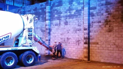 Workers pouring concrete from a cement mixer into a construction site with cinder block walls under blue lighting.