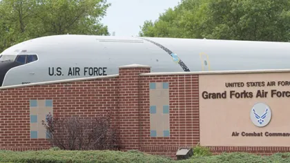 Airplane nose docked behind Grand Forks Air Force Base sign with trees in background