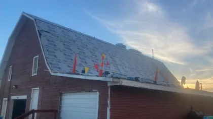 Red barn with roof under construction, covered in waterproofing material, against a blue sky at sunset.