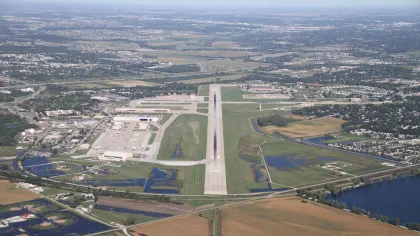 Aerial view of a regional airport with runway, taxiways, parked planes, and surrounding farmland and suburbs.