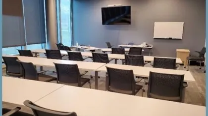 Modern classroom with tables, black mesh chairs, large windows, wall-mounted TV, and whiteboard on blue walls.