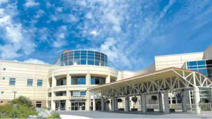Modern hospital building with large glass windows, white facade, and covered entrance under a blue sky with clouds.