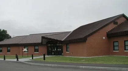 Brick building with a brown tiled roof and glass entrance on a cloudy day with surrounding grass and pavement.