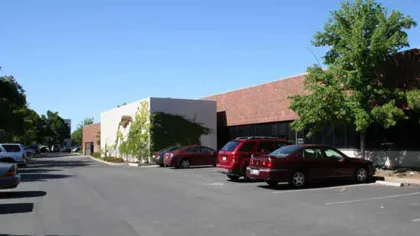 Office building with red brick facade, white walls with ivy, parking lot, and several parked cars under clear blue sky.