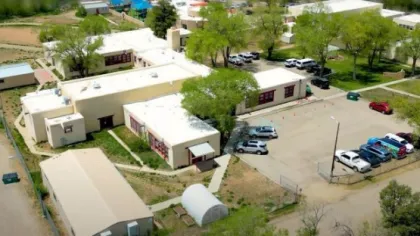 Aerial view of a school campus with white buildings, parking lot, trees, and playground areas under clear skies.